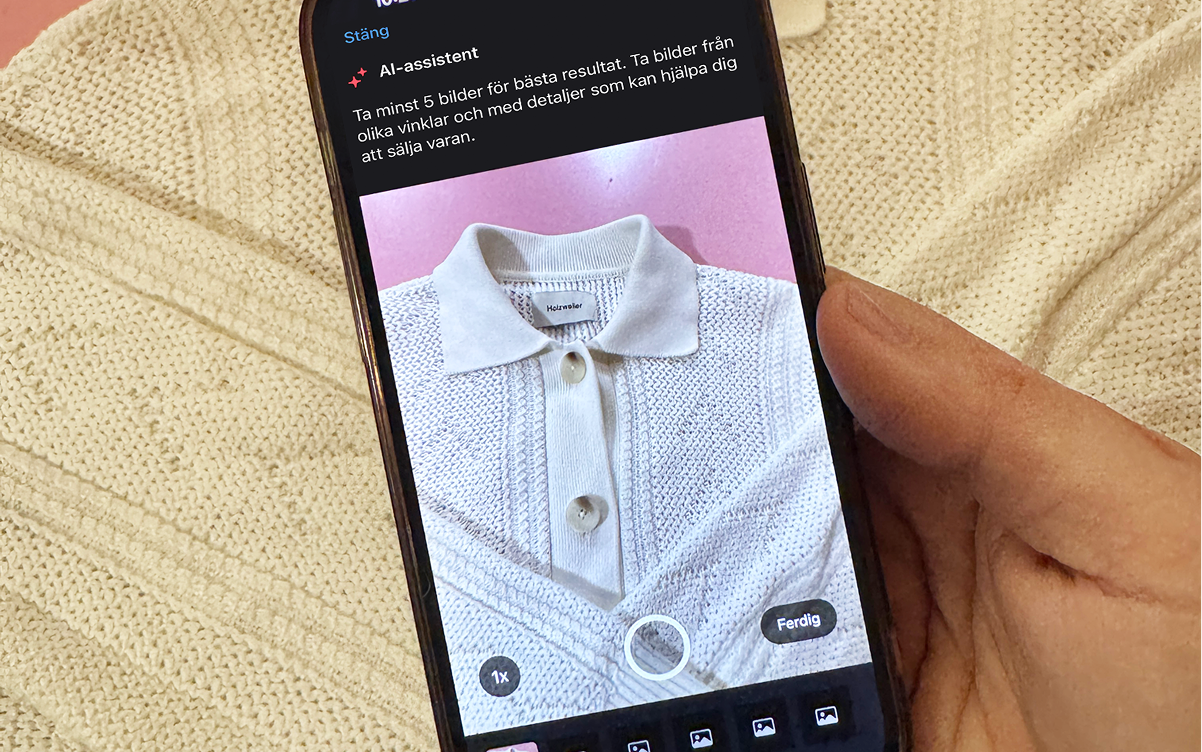 En hand som håller en smartphone som visar en bild på en stickad tröja i off-white mot en rosa bakgrund, med den faktiska tröjan bakom telefonen.