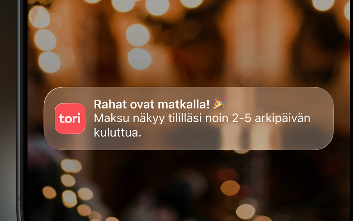 Lähikuva älypuhelimesta, jonka näytöllä näkyy viesti Tori-sovelluksesta, jossa lukee että rahat ovat matkalla ja näkyvät käyttäjän tilillä noin 2–5 arkipäivän kuluessa, sekä taustalla sumea, lämminsävyinen kuva valoista.