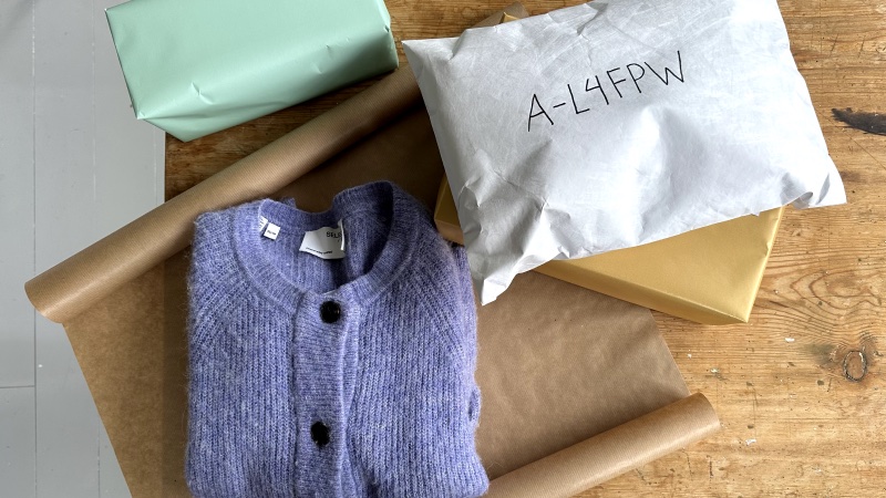 Et brunt bord sett ovenfra, med en grønn pakke og en hvit pakke på toppen, sammen med en lilla genser som skal pakkes inn i brunt papir for frakt.